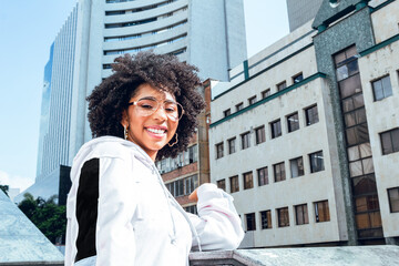 Wall Mural - beautiful afro girl smiling in the city wearing sportswear with beautiful city view of buildings in sunny day outdoors