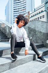 Wall Mural - beautiful afro model girl posing in trendy sportswear in the city sitting on stairs in the city with sneakers and sidadera in sunny day