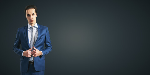 Poster - Young self-made businessman in blue suit isolated on dark grey wall backdrop with empty space for your text or logo, mockup