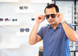 © panyajampatong - Portrait of Indian handsome man looking thoroughly through sunglasses, touching frame of eyewear, wearing a casual outfit,  in an optics store..