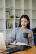 © ArLawKa - A beautiful Asian businesswoman is drinking coffee and working on a laptop with a tiger skin notebook on her desk at the office.
