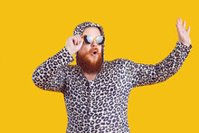 Portrait Of Funny Comic Chubby Man Who Is Strangely Dressed And Having Fun On Orange Background. Cheerful Crazy Millennial Bearded Fat Man Dressed In Leopard Sweater With Hood And Sunglasses.