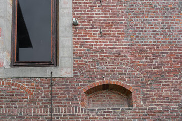 Old brick wall of an ancient red brick building with a blocked niche of a medieval window and a large window next to which there is a security camera and a motion sensor, natural background