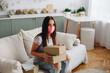 © maxfotoadobe - A girl in a mask sits on a sofa and holds boxes with parcels in her hands in a bright interior waiting for a courier.