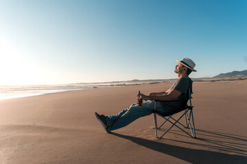 latin man sitting relaxed on the shore of the beach drinking beer and listening to music 