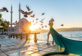 Woman in long green dress posing with flying pigeons at the historical square near Ortaköy mosque and Bosporus waters