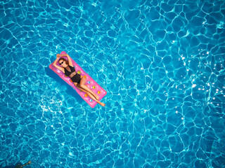 Wall Mural - Fit pretty girl in bikini chilling on inflatable pink mattress in swimming pool. Slim hot woman in swimwear tanning. Female relaxing on float in blue water at luxury resort. Aerial, view from above.