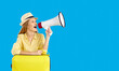 Passenger travel abroad weekends. Woman traveler with megaphone announces interesting offers for tourists on blue background. Woman in hat and with suitcase speaks into loudspeaker near copy space.