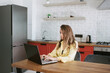 © Марина Демешко - A young beautiful caucasian blonde woman in a yellow shirt sitting at the table working on a laptop from home in the kitchen. Freelance