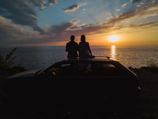 Wall Mural - Silhouette of a happy couple sitting on the roof of a car on a cliff in front of the sea with a charming beautiful orange-blue sky at sunset, view from a drone. Concept of vacations and travel,romance