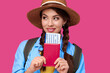 © khmelev - Young female traveler wearing casual clothes and straw hat holding passport with ticket on pink isolate background. Tourism concept. Studio shot