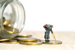 Miniature people toy figure photography. An old men grandfather walking out dragging a cart, from a jar full of coins.
