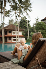 Wall Mural - Young beautiful mother with a baby in her arms sits on a sun lounger. Mom enjoys a moment of tenderness, feeling love for her sweet baby girl. Happy motherhood.