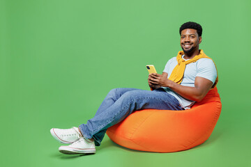Wall Mural - Full size young man of African American ethnicity 20s in blue t-shirt sit in bag chair hold in hand use mobile cell phone isolated on plain green background studio portrait. People lifestyle concept