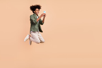 Full length body size view of attractive cheerful guy jumping using device chatting isolated over beige pastel color background