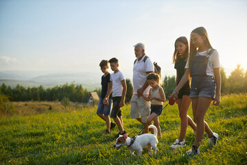 Happy gray man with children walking along mountain hill at summer day. Side view of bearded father with teens and dog backpacking together, with scenic landscape on background. Concept of family.