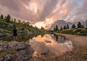 Male hiker gazing at fascinating reflections of the Dolomites mountains in lake Limedes at Sunset. Vivid and bright colors of the dramatic evening.