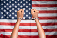 Close Up Shot Of Hanads Rising With Cuffs Against Us Or American Flag - Concept Of Juneteenth, Freedom And Punishment.