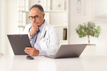 Sticker - Mature male doctor sitting in an office and reading a document  at a desk with a computer