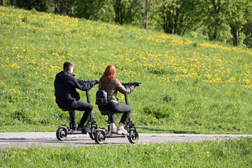 Guy and girl ride electric scooters with seats in summer park on background of green hill with flowers