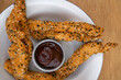 Rustic American food. Top closeup view of fried chicken sticks breaded with sesame seeds and a spicy ketchup sauce, in a white bowl on the wooden table.