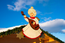 Christmas Angel On A Hut Playing Mandolin. Background Blue Sky And Clouds