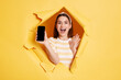 © sementsova321 - Indoor shot of amazed brunette woman stands in torn paper hole, showing cell phone with blank screen for advertisement, looking through breakthrough of yellow background.