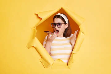 Angry woman in sunglasses and hair band stands in torn paper hole, standing in breakthrough of yellow background, recording voice message with aggressive expression.