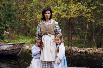 Mother with her two little daughters in vintage dresses in village walking outdoors. Family in retro style clothes standing near lake in park. Portrait of pretty mother and baby girls of 3three and 5