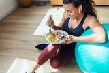 Fototapeta  - Sporty young woman eating healthy while listening to music sitting on the floor at home.