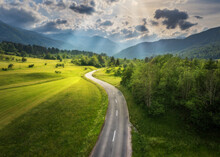 Aerial View Of Road In Green Meadows At Sunset In Summer. Top View From Drone Of Rural Road, Mountains, Forest. Beautiful Landscape With Roadway, Sun Rays, Trees, Hills, Green Grass, Clouds. Slovenia