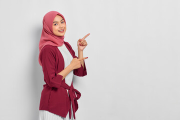 Poster - Cheerful beautiful Asian woman in casual shirt pointing fingers aside at copy space isolated over white background