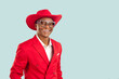 Portrait of happy extravagant dark skinned showman on pastel light blue background. African American man in red jacket, cowboy hat and shiny glasses smiles with snow-white smile near copy space.