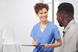 © Mediaphotos - Cheerful Caucasian female doctor sitting on chair in hospital corridor talking to her African American patient