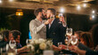 © Gorodenkoff - Big Dinner Party with a Crowd of Multiethnic Diverse Friends Celebrating at a Restaurant. Handsome Happy Gay Hosts Propose a Toast and Raise Wine Glasses while Sitting at a Table in the Evening.