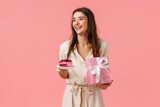 Fototapeta  - Celebration, party and happiness concept. Carefree smiling happy european woman having fun parying own b-day party, holding gift and piece cake, blow-out candle, pink background