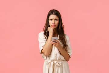 Wall Mural - Sulky cute girlfriend facing complicated decision, frowning and sulking leaning on fist, holding smartphone, read strange message cant understand, feel perplexed, standing pink background