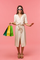 Wall Mural - Full-length vertical portrait elegant good-looking woman on vacation, shopping and having fun in malls, wearing sunglasses, light dress, carry bags from store, standing pink background smiling