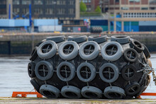 A Large Rubber Boat Bumper On The Edge Of A Dock. Surface Covered With Old Tires Held On By Chains.Water And Buildings In Background.