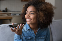 Happy Dreamy Teen African Girl Talking On Mobile Phone On Speaker, Recording Audio Message, Giving Voice Command To Virtual Assistant, Looking Away With Thoughtful Toothy Smile