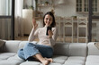 © fizkes - Cheerful happy young millennial woman excited with video call talk, recording self video, waving hand hello at smartphone screen, resting on home sofa indoors, using mobile phone for communication