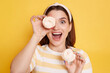 Caucasian excited woman holds marshmallows in hands, wearing striped shirt and glasses, covering her eye with sugary sweets, looking at camera with open mouth, posing isolated over yellow background.
