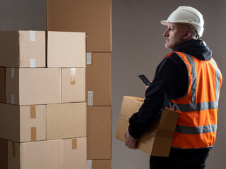 Poster - Warehouse logistics of goods. Man in orange vest next to boxes. Guy works in logistics warehouse. Man with phone looks away. Logistic distribution of parcels. Warehouse employee career.