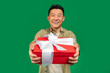 Holiday surprise. Happy middle aged asian man giving gift box to camera and smiling, green studio background