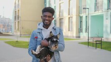 African American Young Man With Smile On Face Embracing Purebred Pug Dog While Standing At Urban Area. Cute Scottish Terrier Relaxing On Owner S Hands After Long Walk On City Street.