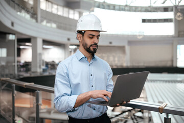 Pensive busy millennial arab guy engineer in safety helmet with beard works with computer controls factory