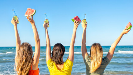 three happy women toasting beers looking at the horizon beach outdoors celebrating summer vacation days. faceless friends raising hands on seaside sunset holidays. joy, freedom, genz and party concept