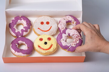 A hand takes a purple-white glazed donut from an orange cardboard box. 6 donuts have 2 buttered smiley face donuts (no holes) 4 purple and white sugar coated donuts sprinkled with crushed dried flower