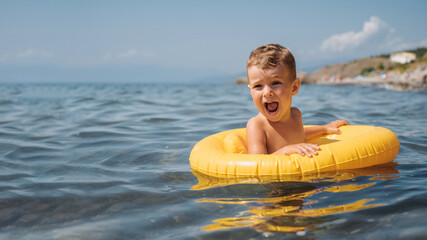 Baby boy swims with an inflatable yellow circle in the sea on a sunny day. The kid learns to swim and enjoys the game.