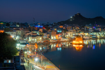 Fototapete - View of famous indian hinduism pilgrimage town sacred holy hindu religious city Pushkar with Brahma temple, aarti ceremony, lake and ghats illuminated at sunset. Rajasthan, India. Horizontal pan